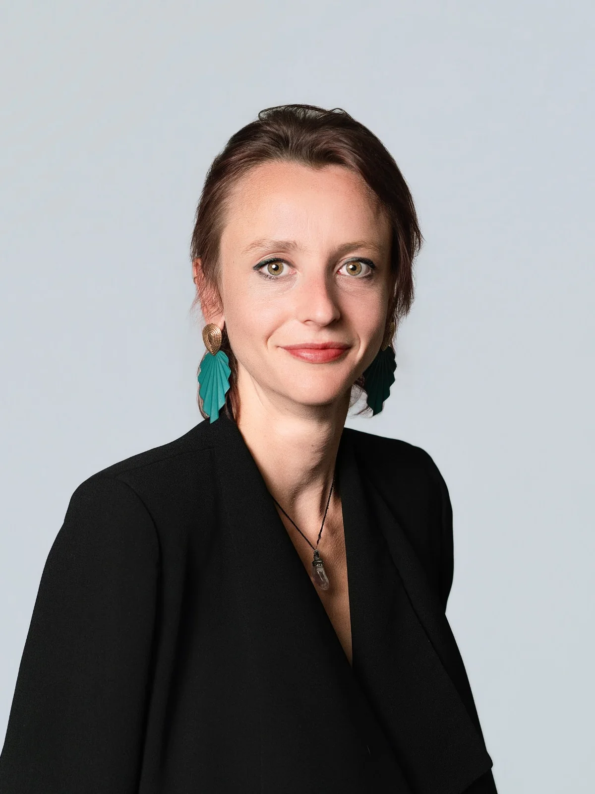 Portrait sur fond gris, en veste noire et boucles d’oreilles vertes.