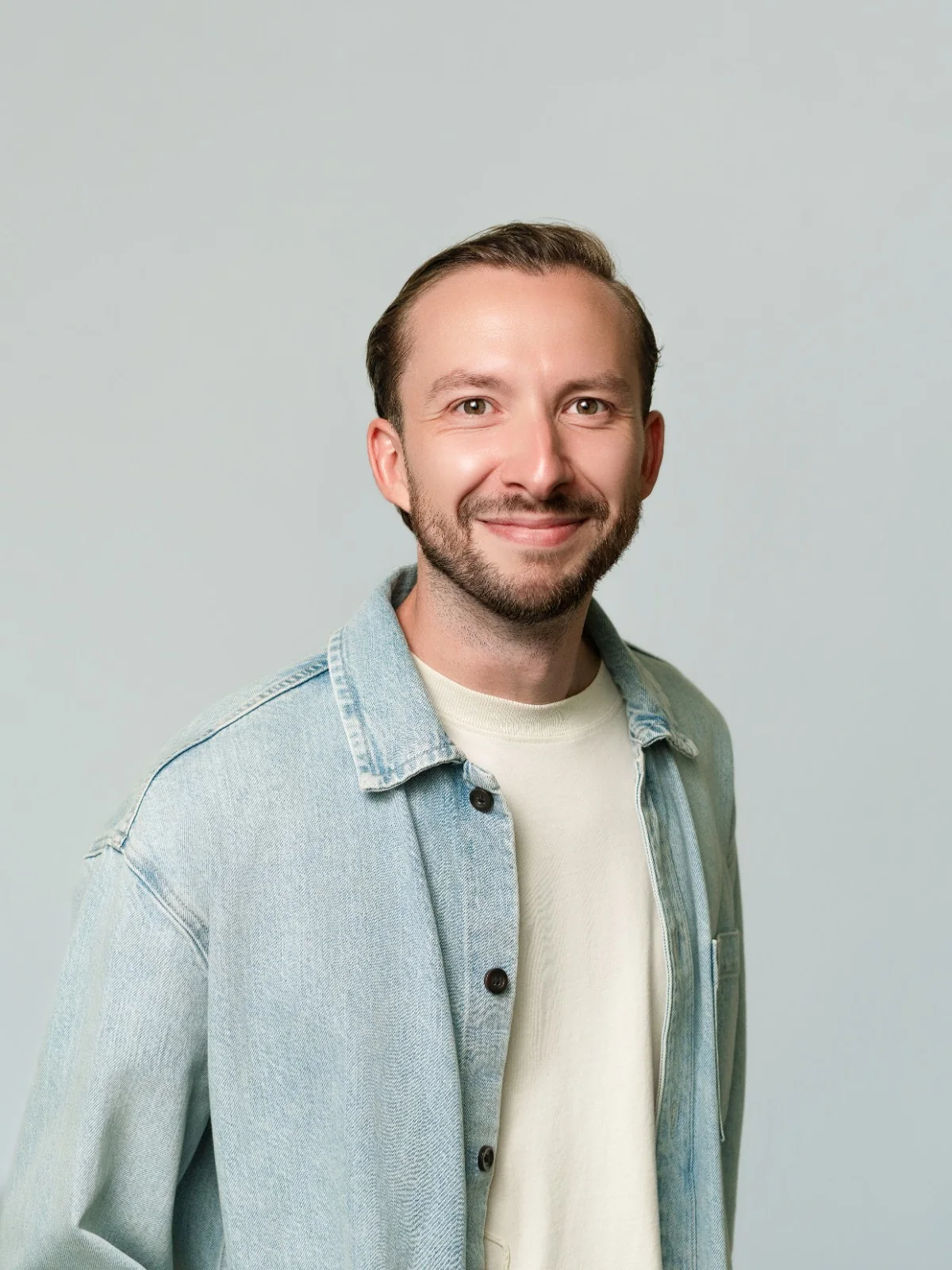 Portrait souriant sur fond gris, en chemise en denim ouverte sur un tee-shirt clair.